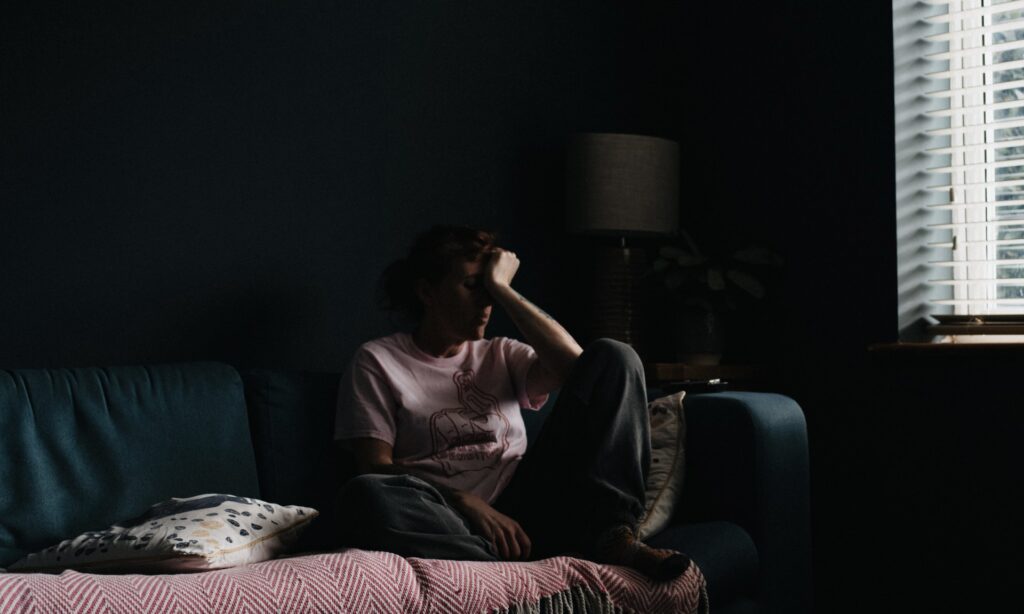 Woman in 30s with brown hair sitting on sofa with face in hands in despair in a dark room | Coercive Control: Spotting the Signs | Irinni Counselling