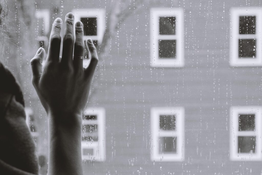 Photo in greyscale of someones hand against a window overlooking another building with rain on the window