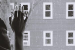 Photo in greyscale of someones hand against a window overlooking another building with rain on the window