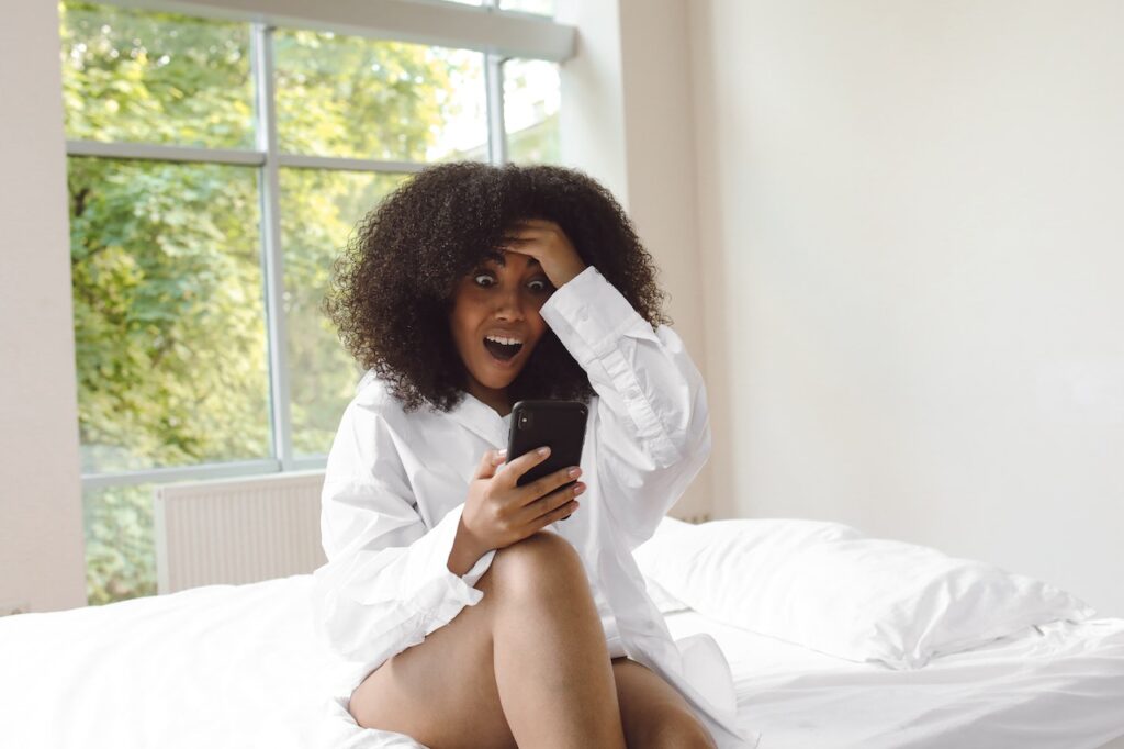 Black woman aged around 35 with afro hair sitting on a bed with a phone in her hand looking surprised about anxiety facts