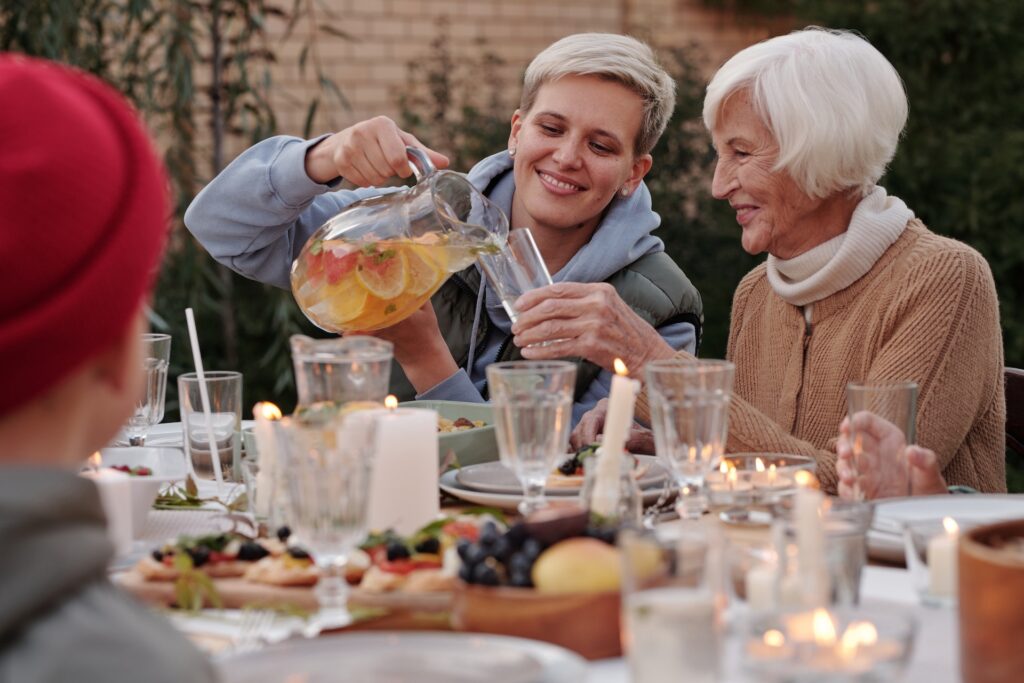 White woman in her 40s with short hair sitting at a table outside, pouring a drink from a jug for an older women with short white hair and a pearl necklace at a family gathering