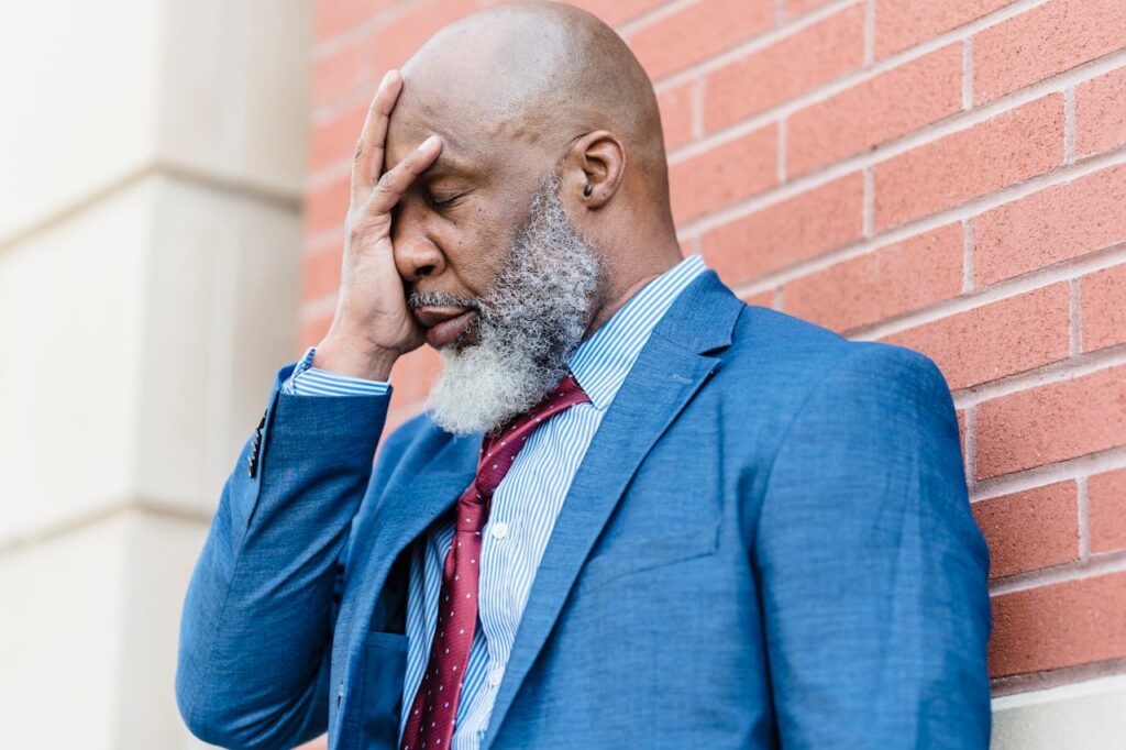 Black man in his 60s experiencing negative self-talk, wearing a blue suit and red tie, leaning on a wall with his head in his hand.