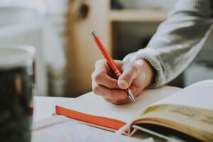 Person holding an orange pen, writing in a notebook.