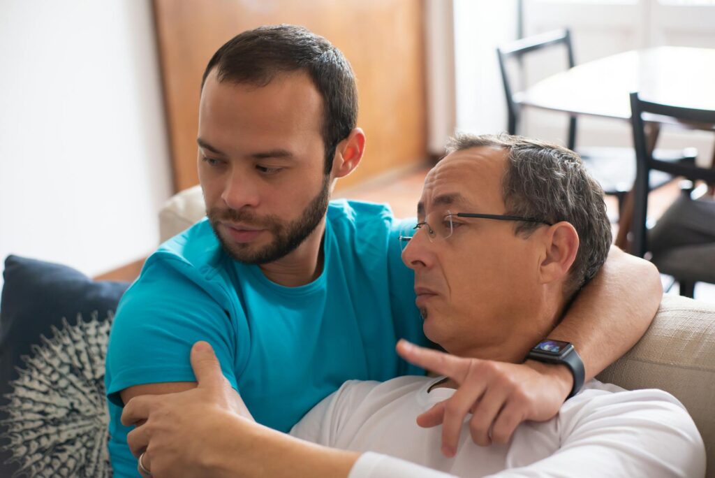 Two men on sofa, younger man in blue tshirt caring for older man in white shirt and glasses, in a caring embrace