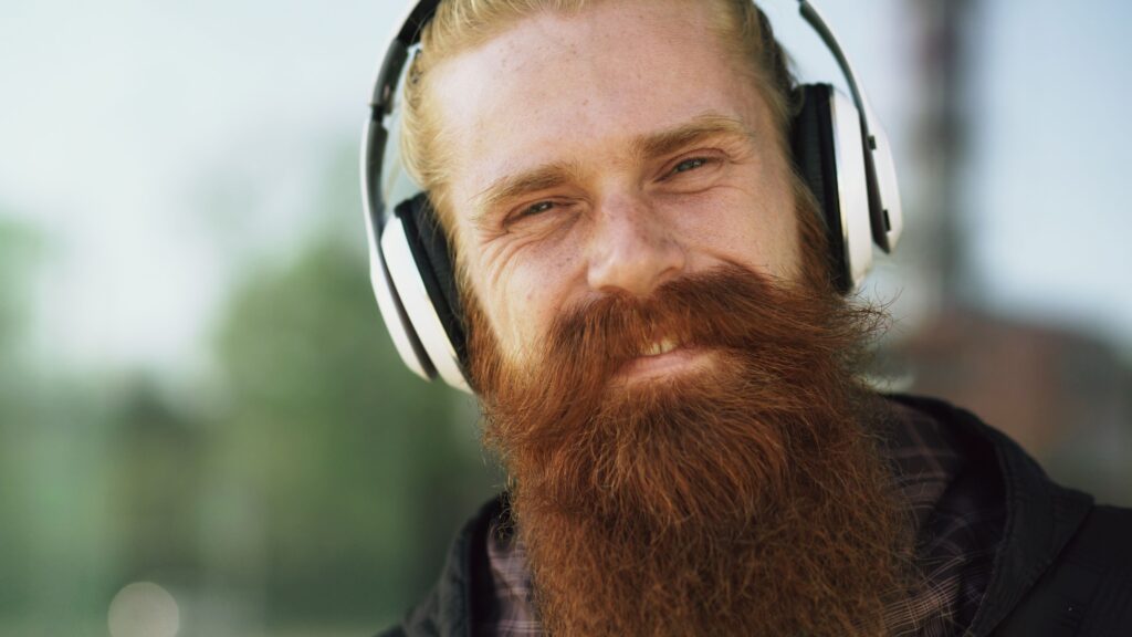 White man with ginger hair and long beard, wearing headphones and smiling