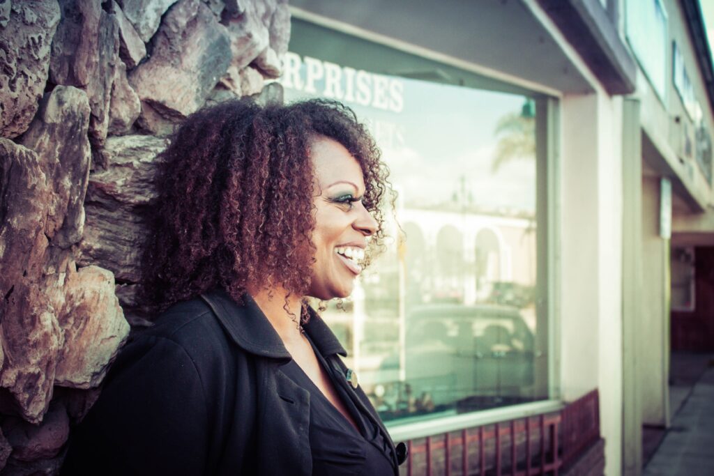 Middle-aged black woman with afro hair standing in the street outside a shop window, with a huge smile and confident look about her